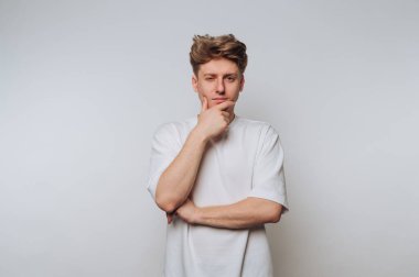 Beyaz tişörtlü genç adam elleri çenesinde gri arka planda düşünüyor..