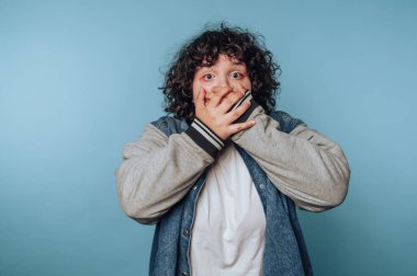 Kıvırcık saçlı biri şaşırmış görünüyor. İki elini de mavi arka planda tutuyor..