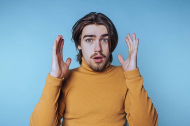 Sarı balıkçı yaka kazak giyen, uzun saçlı, şaşırmış genç bir adam açık mavi bir arka plana karşı ellerini kaldırıyor.
