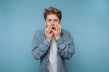 Kot gömlekli genç adam mavi arka planda şok olmuş görünüyor..