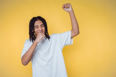 Saçları örgülü genç adam sarı arka planda kutlama yapıyor..