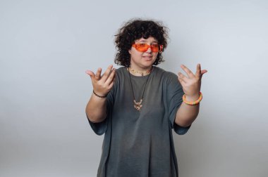 Kıvırcık saçlı, turuncu güneş gözlüğü takan ve sıradan kıyafetler giyen, elleri arka planda olan biri..