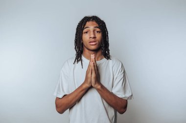 Saçlarını örmüş beyaz tişörtlü, elleri dua pozisyonunda genç bir adam..