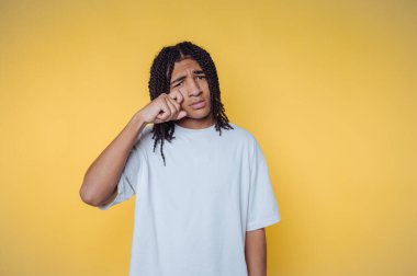 Saçları örgülü, sarı arka planda gözyaşları silen bir genç..