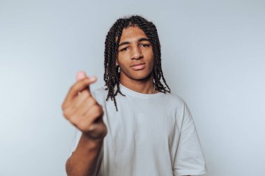 Saçları örgülü genç bir adam düz bir arkaplanda el hareketi yapıyor..