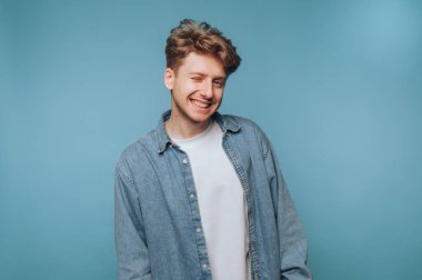 Kot gömlekli genç adam mavi arka planda göz kırpıyor ve gülümsüyor..
