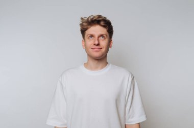 Beyaz tişörtlü genç adam arka planda yukarıya bakıyor..