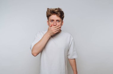 Beyaz tişörtlü, ağzı elle kaplı genç bir adam, şaşırmış bir ifade..
