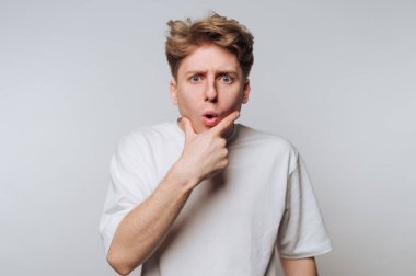 Beyaz tişörtlü genç adam şaşırmış bir ifadeyle, elleri çenesinde.