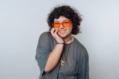 Kıvırcık saçlı, turuncu güneş gözlüklü ve gri tişörtlü gülümseyen biri..