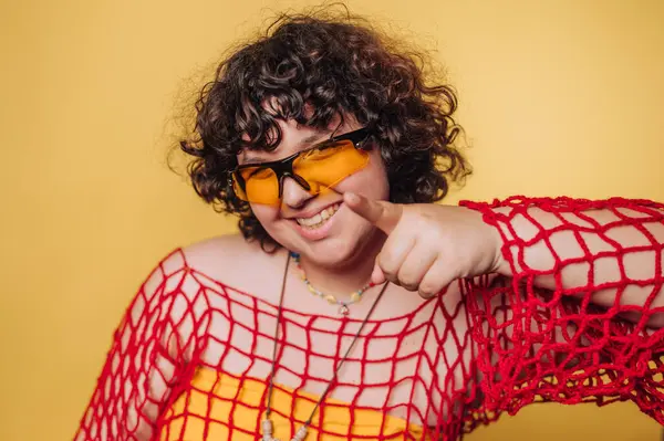Kıvırcık saçlı, sarı güneş gözlüğü takan ve sarı arka planda kırmızı ağ takan gülümseyen biri..