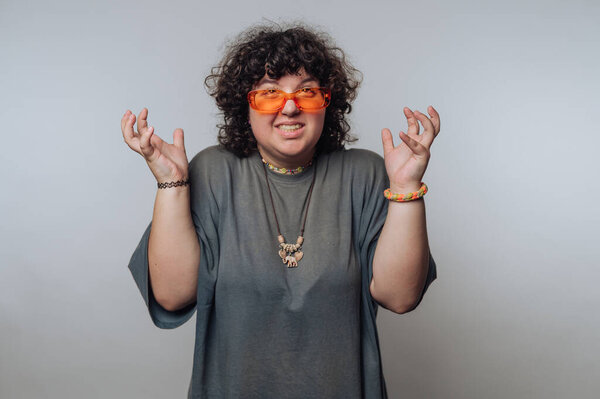 Человек с вьющимися волосами в оранжевых солнцезащитных очках и серой рубашке, жестикулирующий с поднятыми руками, на обычном фоне.