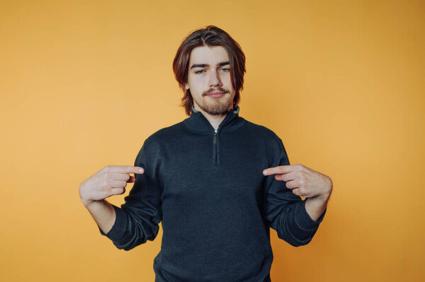 Молодой человек в темном свитере указывает на себя на оранжевом фоне.