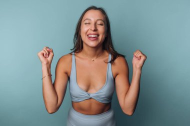 Açık mavi spor kıyafetli neşeli genç bir kadın pastel mavi arka planda yumruklarını kaldırarak kutluyor.