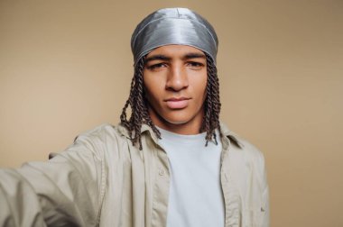 Saçları örgülü, gümüş rengi uzun ve bej gömlekli genç bir adam..