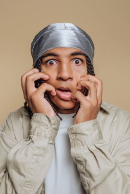 Gri bir başörtüsü ve bej ceket giyen, tarafsız bir arkaplan karşısında şaşırmış bir genç..