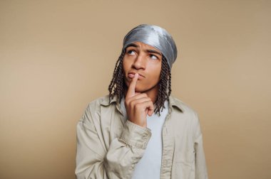 Saçları örgülü, gümüş bir başörtüsü takan bej bir arka planda düşünceli bir genç..