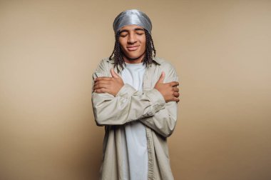 Örgülü saçlı, kendine sarılan, başörtüsü takan, bej arka planda genç bir adam..