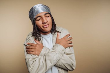 Saçları örgülü, bej gömlekli genç bir adam. Tarafsız bir arkaplan karşısında sakin bir ifadeyle kendine sarılıyor..