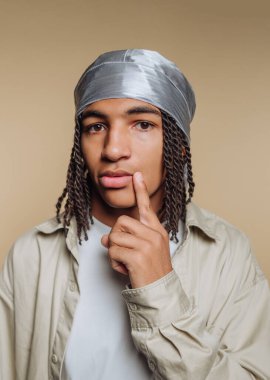 Saçları örgülü, gümüş bir durak giyen genç bir adam bej arka planda düşünceli bir şekilde poz veriyor..