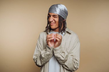 Saçları örgülü genç adam bandana takıyor, göz kırpıyor ve gülümsüyor..