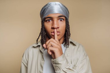 Saçları örgülü, gümüş bir durag takan genç bir adam. Bej bir arka planda, parmakları dudaklarında bir şşş hareketi yapıyor..