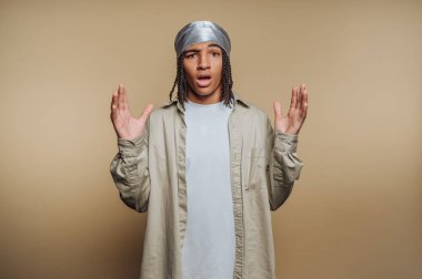 Şaşırmış bir ifadeyle genç bir adam, Durag ve günlük kıyafetler giyiyor, bej bir arka plana karşı duruyor..