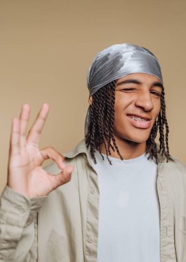 Örgülü genç adam gümüş bir durag takıyor, göz kırpıyor ve bej bir arka plana 