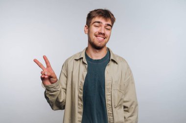 Genç adam gülümsüyor ve gri arka planda barış işareti gösteriyor..