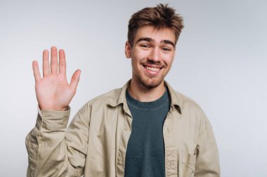 Genç adam gülümseyip arka planda kameraya el sallıyor..