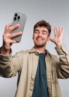 Genç adam akıllı telefonuyla selfie çekiyor, gülümsüyor ve el sallıyor..