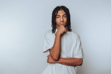 Sade bir arka plana karşı düşünceli bir duruşta örgülü saçlı genç bir adam..