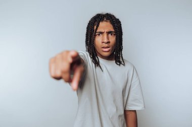 Saçları örgülü genç bir insan kamerayı işaret ederek ciddi ya da agresif bir duyguyu ifade eder. Arkaplan sade ve açık renkli, konuyu vurguluyor..