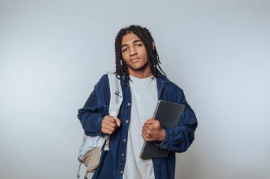 Saçları örgülü genç bir adam dizüstü bilgisayar ve sırt çantası tutuyor.