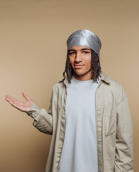 A young person with braided hair wearing a silver headscarf and a light-colored shirt, posing against a beige background