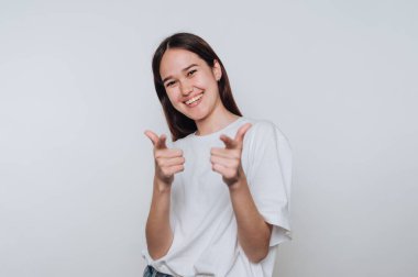A young woman stands against a plain wall, smiling brightly. She poses with her hands, giving a thumbs up while wearing a simple white t-shirt and looking cheerful.