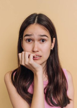 A young woman shows a worried expression, biting her nails in a relaxed indoor environment. The soft yellow background adds to the emotional intensity of the moment.