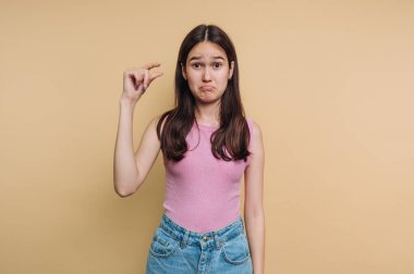A young woman stands in front of a beige wall, showing a disappointed expression while holding up a small amount with her fingers. She wears a pink top and denim jeans.
