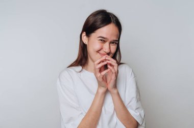 A woman is smiling brightly while holding her hands together near her face. Her playful expression hints at amusement or a fun secret. The background is simple and light-colored.