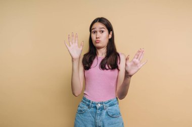 A young woman stands against a soft beige backdrop. She raises her hands in surprise while making a playful expression. Her casual outfit features a pink top and denim shorts.