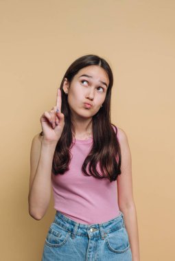 A young girl stands against a solid background with a playful expression. She raises her finger as if to indicate a thought. Her casual outfit adds to the relaxed vibe of the moment.