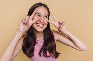 A young girl smiles brightly while making peace signs with her fingers. She has long hair and wears a pink top, enjoying a fun moment against a soft yellow backdrop.