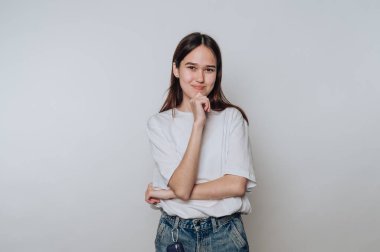 A young woman poses thoughtfully in a simple outfit, with her hand resting on her chin. She stands confidently against a plain background in a relaxed atmosphere.