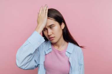 A young woman stands in a softly lit pink room, expressing frustration with her hand on her forehead. Her emotions reflect tiredness and stress, revealing a relatable moment.