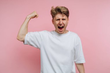 A young man in a white t-shirt stands with a raised fist and a fierce expression. He appears to be expressing strength and determination in front of a soft pink backdrop.