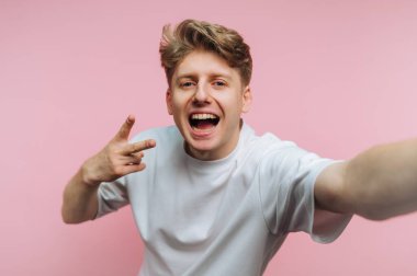 A cheerful young man poses with a wide smile and peace sign in front of a bright pink backdrop, capturing a fun and playful moment filled with joy.