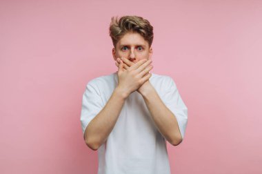 A young man stands against a pink background, covering his mouth with both hands. His expression reveals surprise or shock, capturing a moment of creativity and emotion.
