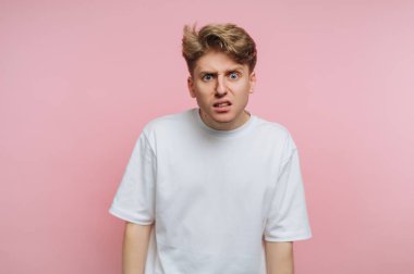 A young man stands with a puzzled look on his face. He is wearing a plain white t-shirt. The background is a soft pink color, creating a vibrant atmosphere.