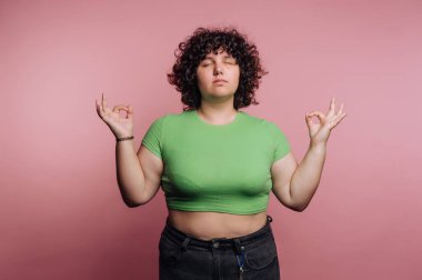 A young woman with curly hair is practicing meditation with her eyes closed. She wears a green top and holds her hands in a relaxed pose, promoting inner peace against a pink backdrop.
