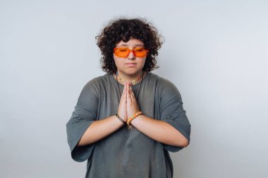 A person stands with hands pressed together in a thoughtful pose, wearing orange sunglasses and comfortable clothing in a serene setting. The atmosphere feels peaceful and focused.